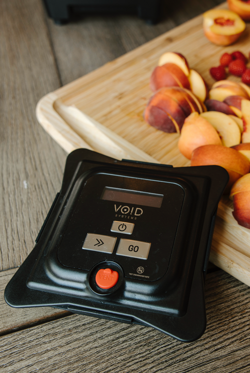 The VOID vacuum lid on a wooden table beside sliced peaches and strawberries