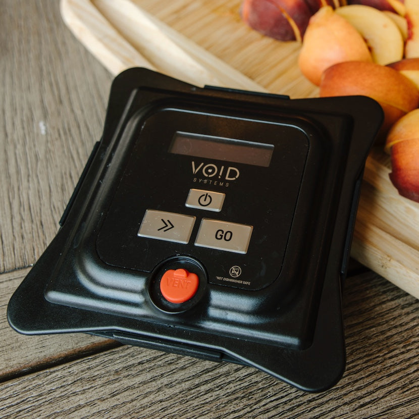 The VOID vacuum lid on a wooden counter next to a cutting board of sliced peaches