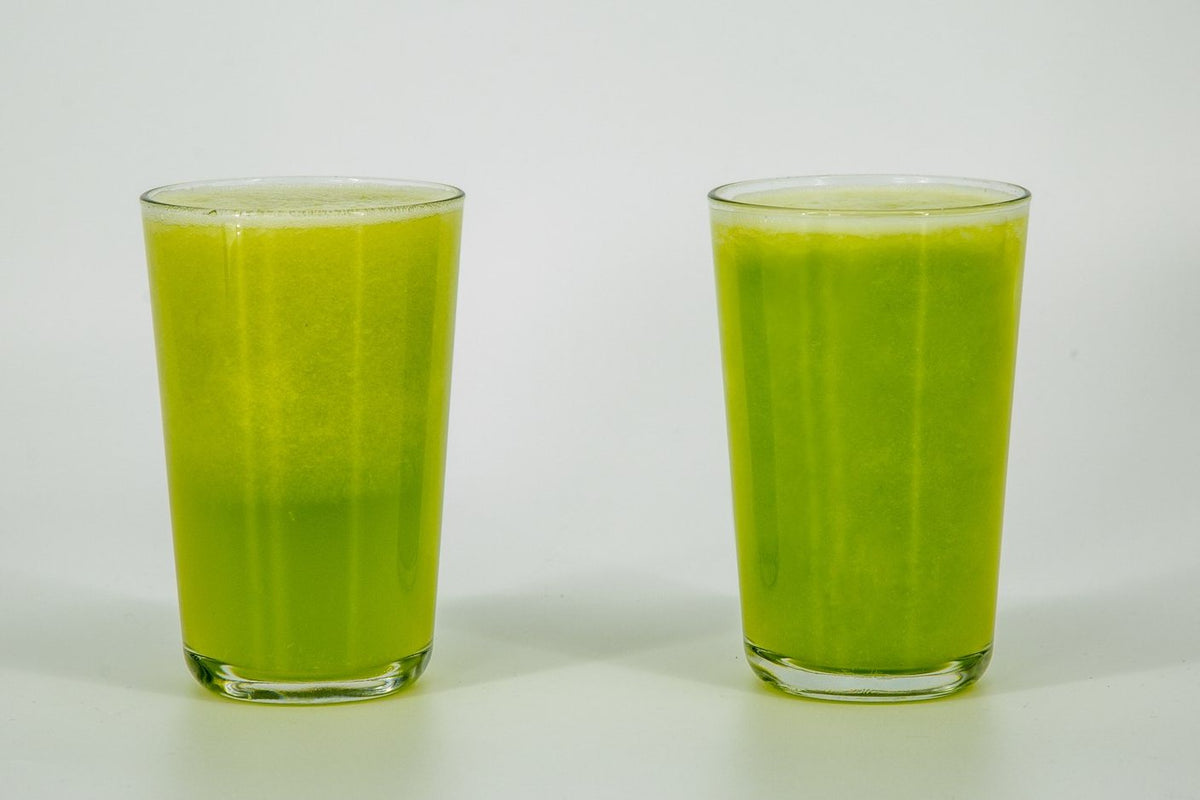 Two glasses of celery juice side by side. The air-blended glass on the left has shifted yellow-green; the VOID glass on the right holds a deeper green.